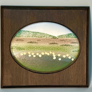 May include: A square dark brown wooden frame containing an oval-shaped enamel plaque. The plaque features a landscape scene with green hills, a textured tan field, and a foreground of green grass with small white circular flower-like shapes.