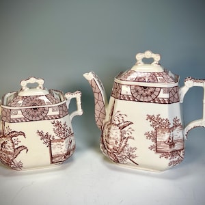 May include: Two-piece antique tea set with a cream-colored base and a maroon transferware design. The set includes a teapot and a sugar bowl, both with lids and ornate handles. The design features landscape scenes and geometric patterns.