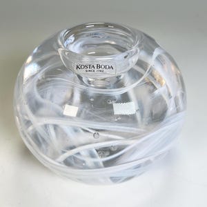 May include: A clear glass votive candle holder with white swirling patterns. The candle holder has a round base and a small, circular opening at the top. The text "KOSTA BODA SINCE 1742" is printed on the side.