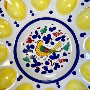 May include: A white ceramic egg serving platter featuring a central hand-painted illustration of a yellow bird surrounded by blue and green floral motifs. The outer rim contains twelve circular indentations, each painted with a yellow egg design.