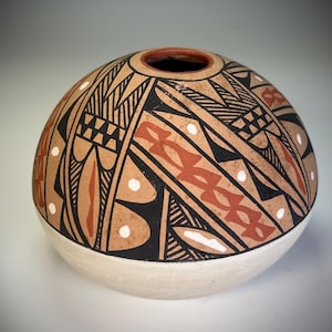 May include: A handcrafted, round ceramic pot with a cream-colored base and a tan upper section. The upper section features intricate geometric patterns in black, red, and white. The pot has a small opening at the top and is likely used for decorative purposes.
