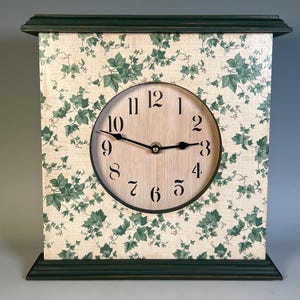 May include: A wooden mantel clock with a light wood clock face and black numerals. The clock's face is surrounded by a cream-colored background with a green ivy pattern. The clock's frame is dark green.