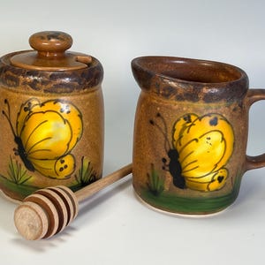 May include: A ceramic honey pot with a lid and a creamer, both decorated with a yellow butterfly design. A wooden honey dipper rests in front. The set has a brown and green color scheme.