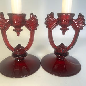 May include: Two ruby red glass candle holders with a decorative design. Each holder features a round base, a central cup for the candle, and a curved, openwork design. The candle holders are a classic and elegant home decor item.