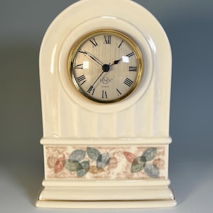 May include: Cream-colored Lenox quartz clock with a gold-tone bezel and Roman numeral hour markers. The clock features a decorative floral band and a vertical striped design. The clock is a decorative home decor item.