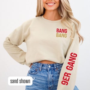 Könnte beinhalten: Ein beiger Pullover mit den Worten "Bang Bang" auf der Vorderseite und "9er Gang" auf dem rechten Ärmel in Rot. Das Model trägt eine blaue Jeans.