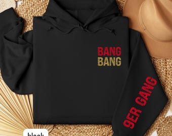 Op de afbeelding: Zwarte hoodie met de tekst "BANG BANG" in rood en goud op de borst. De mouw heeft de tekst "9ER GANG" in rood. Het woord "black" staat in een witte doos onderaan.