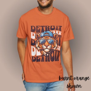 May include: Burnt orange t-shirt with a graphic of a tiger wearing sunglasses and a baseball cap. The text "DETROIT" is repeated in a layered, distressed style.