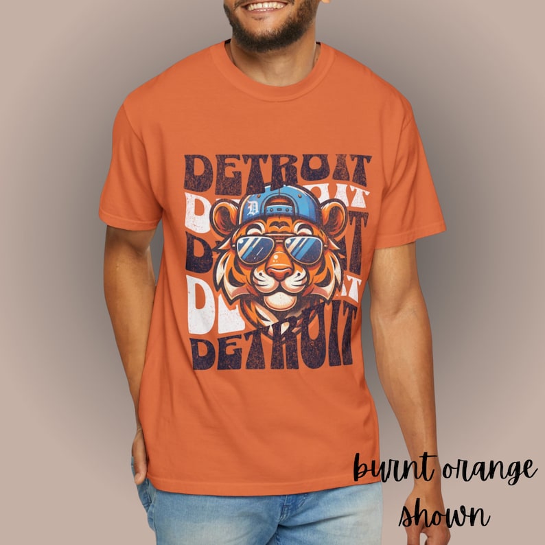 May include: Burnt orange t-shirt with a graphic of a tiger wearing sunglasses and a baseball cap. The text "DETROIT" is repeated in a layered, distressed style.