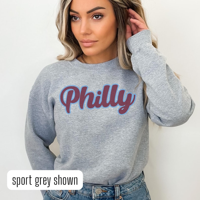 May include: A grey sweatshirt with the word "Philly" in a retro font in maroon and blue. The model is wearing the sweatshirt and has long brown hair. The text "sport grey shown" is at the bottom of the image.
