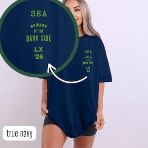 以下が含まれることがあります： 紺色のTシャツ。前面には、緑色で「SEA BEWARE OF THE DARK SIDE LX '26」と書かれています。Tシャツは無地で、ゆったりとしたフィット感です。文字はTシャツに縦に並んでいます。