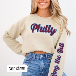 May include: A beige sweatshirt with the word "Philly" in maroon and blue lettering on the front and the phrase "Ring the Bell" in maroon and blue lettering on the sleeve. The model is wearing blue jeans.