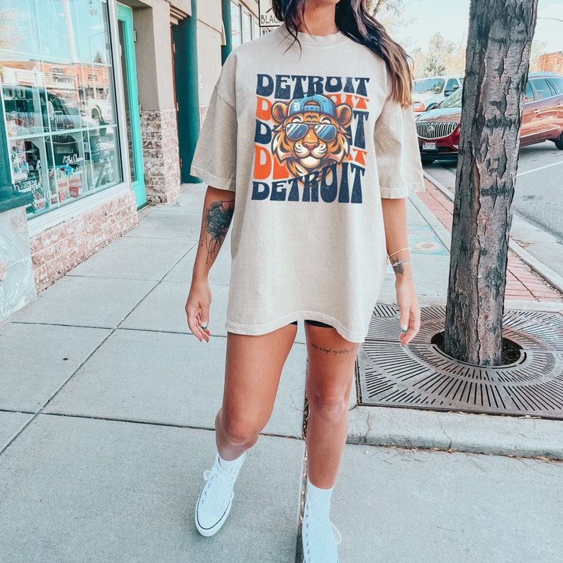 May include: A woman wearing a beige oversized t-shirt with a graphic print of a tiger wearing sunglasses and a baseball cap. The graphic is repeated in a layered effect with the word "DETROIT" printed in black and orange. The t-shirt is a loose fit and has short sleeves.