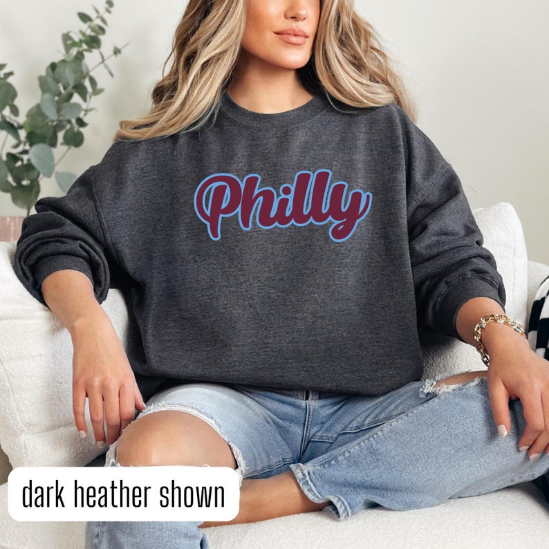 May include: A dark heather grey sweatshirt with the word "Philly" in a maroon and blue script font.
