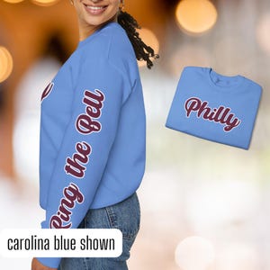 May include: A light blue sweatshirt with the phrase "Ring the Bell" printed in maroon on the left sleeve. The sweatshirt is shown on a model. A separate light blue t-shirt with the word "Philly" printed in maroon is also shown.