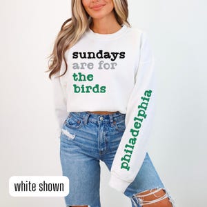 Może przedstawiać: Biała bluza z napisem "Sundays are for the birds" z przodu i "Philadelphia" na rękawie. Modelka nosi niebieskie dżinsy.