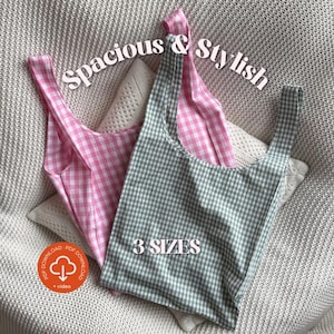 May include: Two gingham-patterned tote bags, one pink and one green, are displayed on a textured surface. The bags have a spacious design and are labeled "Spacious & Stylish" and "3 Sizes". The bags are made of fabric.
