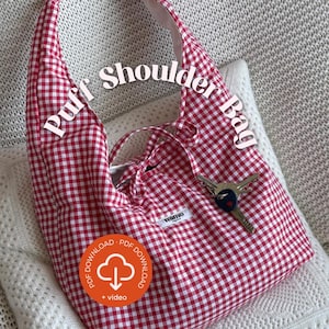 Może przedstawiać: Czerwono-biała torba na ramię w kratę z napisem "Puff Shoulder Bag". Torba ma miękki, puszysty design i zapięcie na sznurek. Klucze są przymocowane do torby. Torba leży na białej poduszce.