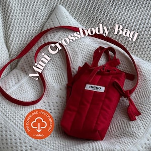 Könnte beinhalten: Eine rote Mini-Umhängetasche mit langem Riemen, auf einer weißen, strukturierten Oberfläche. Die Tasche hat eine rechteckige Form mit vertikalen Nähten und ein kleines weißes Etikett mit der Aufschrift "mimo". Der Text "Mini Crossbody Bag" steht oben.