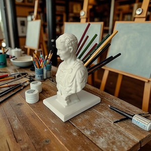 May include: A white plaster bust of a classical figure sits on a white base on a rustic wooden table. Paintbrushes, pencils, and art supplies are arranged around the bust. Easels with blank canvases are in the background, suggesting an art studio.