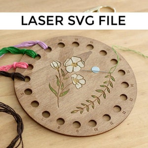 May include: A wooden embroidery floss organiser with a floral design and the text "LASER SVG FILE". The organiser has multiple holes for holding floss and is decorated with a floral design and a leafy branch. Various colours of embroidery floss are visible.
