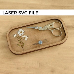 Puede incluir: Una bandeja de madera con un diseño floral y un par de tijeras pequeñas. La bandeja también tiene un pequeño círculo con tres agujas.