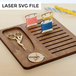 Puede incluir: Un organizador de hilo de bordar de madera con ranuras para carretes de hilo, un pequeño compartimento para tijeras y un compartimento circular para agujas. El organizador está hecho de madera marrón oscura y tiene un acabado de madera natural. El organizador está sobre una superficie marrón claro.