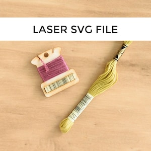 May include: A wooden thread bobbin with pink thread wound around it. The bobbin has a label with the number "312" printed on it. A yellow embroidery floss with the number "734" printed on the label is lying on a wooden surface.