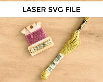 May include: A wooden thread bobbin with pink thread wound around it. The bobbin has a label with the number "312" printed on it. A yellow embroidery floss with the number "734" printed on the label is lying on a wooden surface.