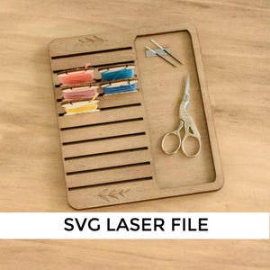 Peut inclure: Organisateur en bois avec des fentes pour ranger le fil à broder et une petite paire de ciseaux. L'organisateur a un motif floral et le texte "SVG Laser File" est imprimé en dessous.