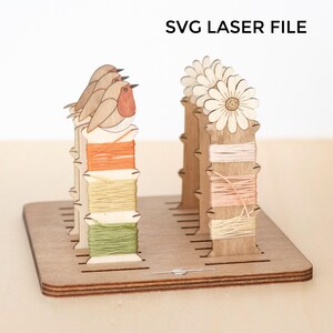 May include: Wooden thread organizer with laser-cut bird and flower designs. The organizer holds spools of colorful thread in orange, yellow, green, and pink. The text "SVG LASER FILE" is at the top.