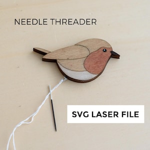 Puede incluir: Un enhebrador de agujas de madera con forma de pájaro con pecho rojo, alas marrones y pico negro. Se muestran los textos "NEEDLE THREADER" y "SVG LASER FILE". Una aguja e hilo blanco están unidos.