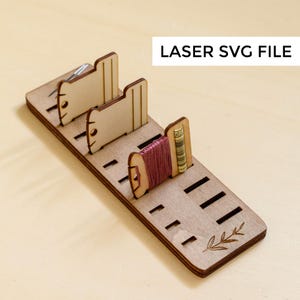 May include: Wooden thread organizer with slots for spools of thread and a small leaf design. The organizer is made of light brown wood and has a label that says "LASER SVG FILE".