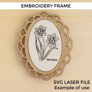 Puede incluir: Marco de bordado de madera con borde festoneado cortado con láser. El centro ovalado presenta un inserto de tela con un dibujo lineal negro de dos narcisos y la palabra "Narcissus". El texto "EMBROIDERY FRAME" está en la parte superior.