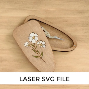 Puede incluir: Una caja de madera con un diseño floral y un pequeño par de tijeras en su interior. La caja está hecha de madera marrón claro y tiene una tapa con bisagras. El diseño floral es en blanco y verde y presenta tres flores. Las tijeras son plateadas y tienen un mango en forma de pájaro. LASER SVG FILE