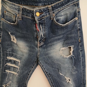 May include: Blue denim jeans with distressed details, including rips and patches. The jeans have a button closure and a red tag near the zipper. The denim is faded in areas, giving a worn appearance.