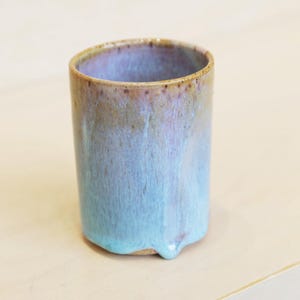 May include: A handmade ceramic tumbler with a unique glaze. The cup features a gradient of colors, transitioning from a brown rim to a light blue base. The bottom has a distinctive drip design. This artful piece is perfect for beverages.