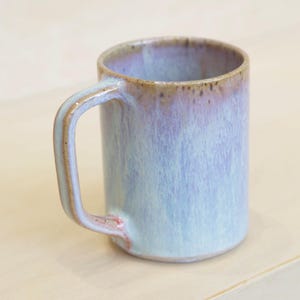 Puede incluir: Una taza de cerámica con forma cilíndrica y un asa curva. La taza presenta un degradado de colores, pasando del azul claro en la base a un tono púrpura en el borde. El asa y el borde tienen un acento marrón.