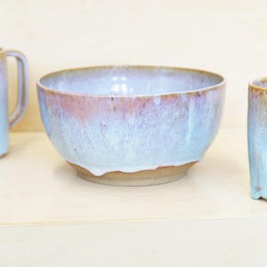 Könnte beinhalten: Eine Keramikschale mit hellblauer und rosa Glasur. Die Schale hat eine breite, abgerundete Form und steht auf einem natürlichen, unglasierten Sockel. Die Glasur tropft an den Seiten herunter und erzeugt eine einzigartige Textur. Eine Tasse ist links zu sehen.