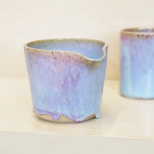 Puede incluir: Dos tazas de cerámica con un esmalte azul claro y morado. La taza de la izquierda tiene un pico y un borde ligeramente irregular. El esmalte gotea por los lados, creando una textura única. Las tazas están sobre una superficie clara.