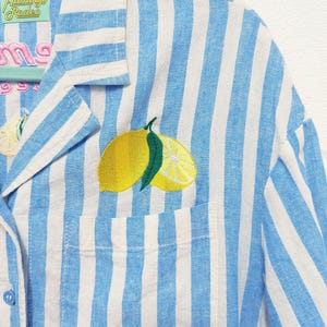 May include: A blue and white striped shirt featuring an embroidered lemon design on the pocket. The shirt has a button-down front and a collar. The text "Flamingo Rodeo" is on a tag near the collar. The word "some" is embroidered on the shirt.