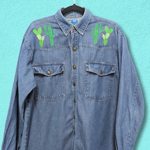 May include: A blue denim shirt with embroidered cactus designs on the chest. The shirt has a button-down front and two chest pockets.