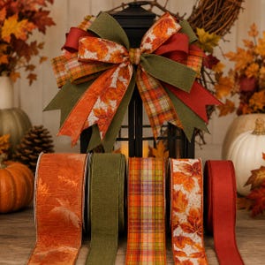 May include: A decorative bow featuring a mix of olive green, solid red, plaid, and orange maple leaf patterned ribbons, displayed atop a black lantern. Below, five individual spools of matching ribbon are arranged in a row on a wooden surface.