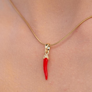 May include: A gold necklace featuring a small, red chilli pepper pendant. The pendant has a gold top with small, clear embellishments. The necklace is worn against a white top.