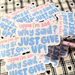 May include: A collection of white stickers with a cloud-like shape. The stickers have the phrases "Oppa I'm sad," "Why sad?" and "Just give up!" in pink and blue bubble letters. A pencil is visible in the background.