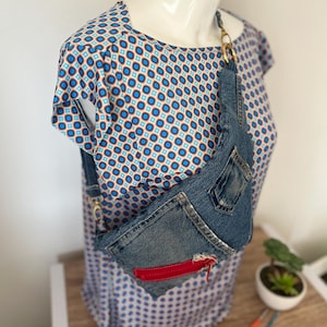 Könnte beinhalten: Eine Jeans-Bauchtasche mit rotem Akzent und goldfarbener Hardware. Die Tasche besteht aus recyceltem Jeansstoff und hat einen Used-Look. Die Tasche wird auf einem Schaufensterpuppe getragen, die ein blau-weiß gemustertes Oberteil trägt.