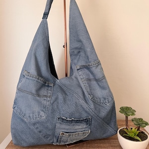 May include: A light blue denim tote bag with multiple pockets, including two large back pockets and a smaller front pocket. The bag has a wide shoulder strap and is made from recycled denim. A small succulent plant in a white pot sits nearby.