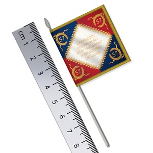 May include: A small, decorative flag with a silver pole. The flag is rectangular, featuring red, blue, and white sections with gold trim. It has the number "57" in multiple places and French text. A ruler is placed next to the flag, showing measurements in centimeters.