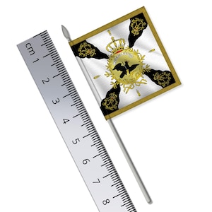 Puede incluir: Una pequeña bandera blanca cuadrada con un borde dorado y un emblema detallado con una corona, un águila y texto. La bandera está montada en un palo plateado. Una regla muestra que la bandera mide aproximadamente 5 cm de largo.