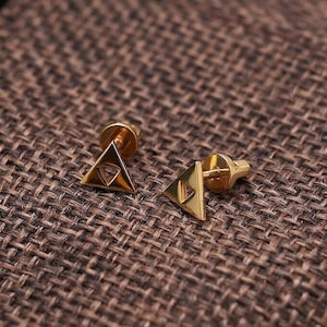 Könnte beinhalten: Ein Paar goldfarbene Dreiecksohrringe mit geometrischem Design. Die Ohrringe liegen auf einem braunen Jutesäckchen.